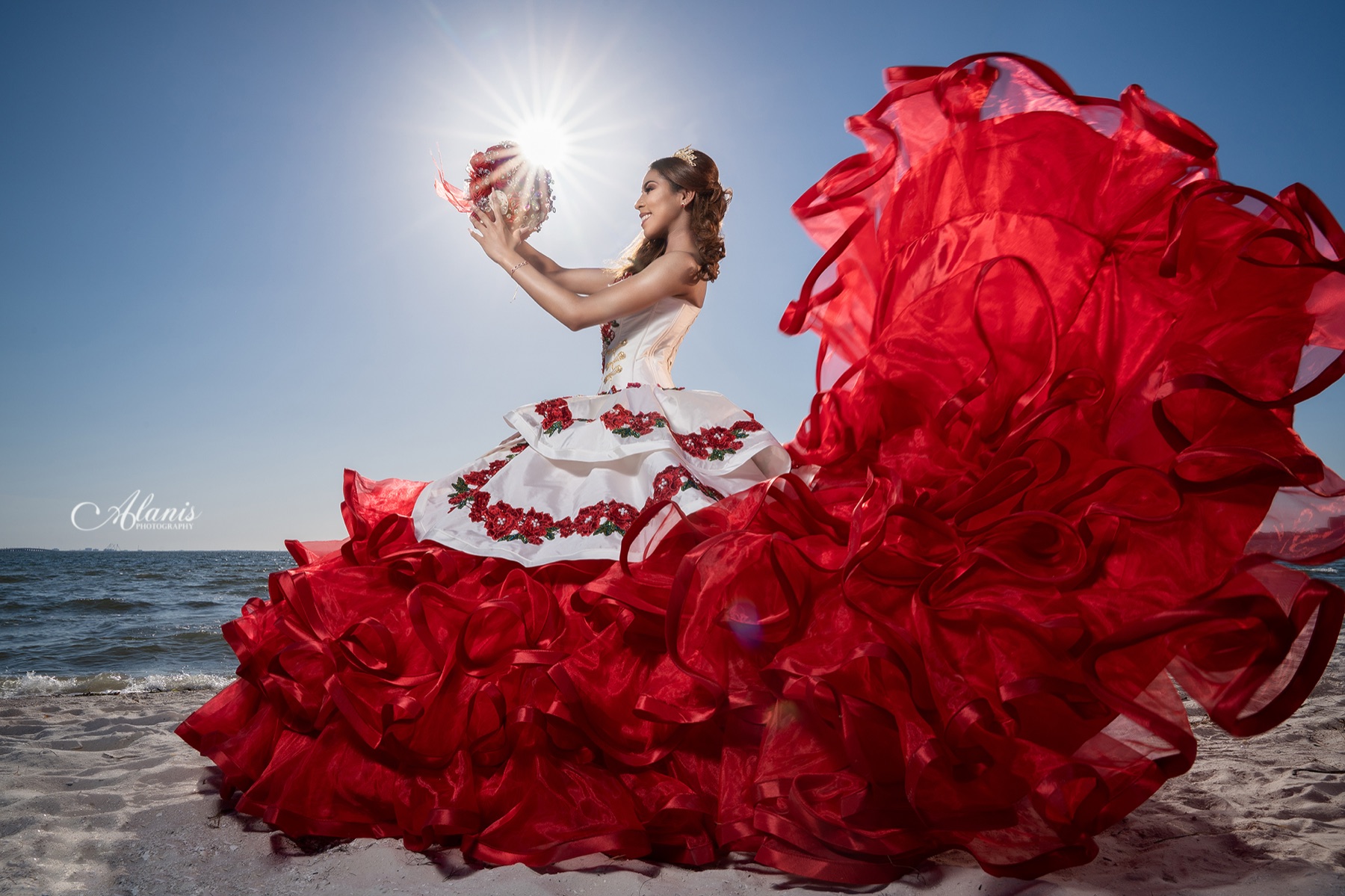 Quinceañera in a red and white gown with its train flying beside the sea